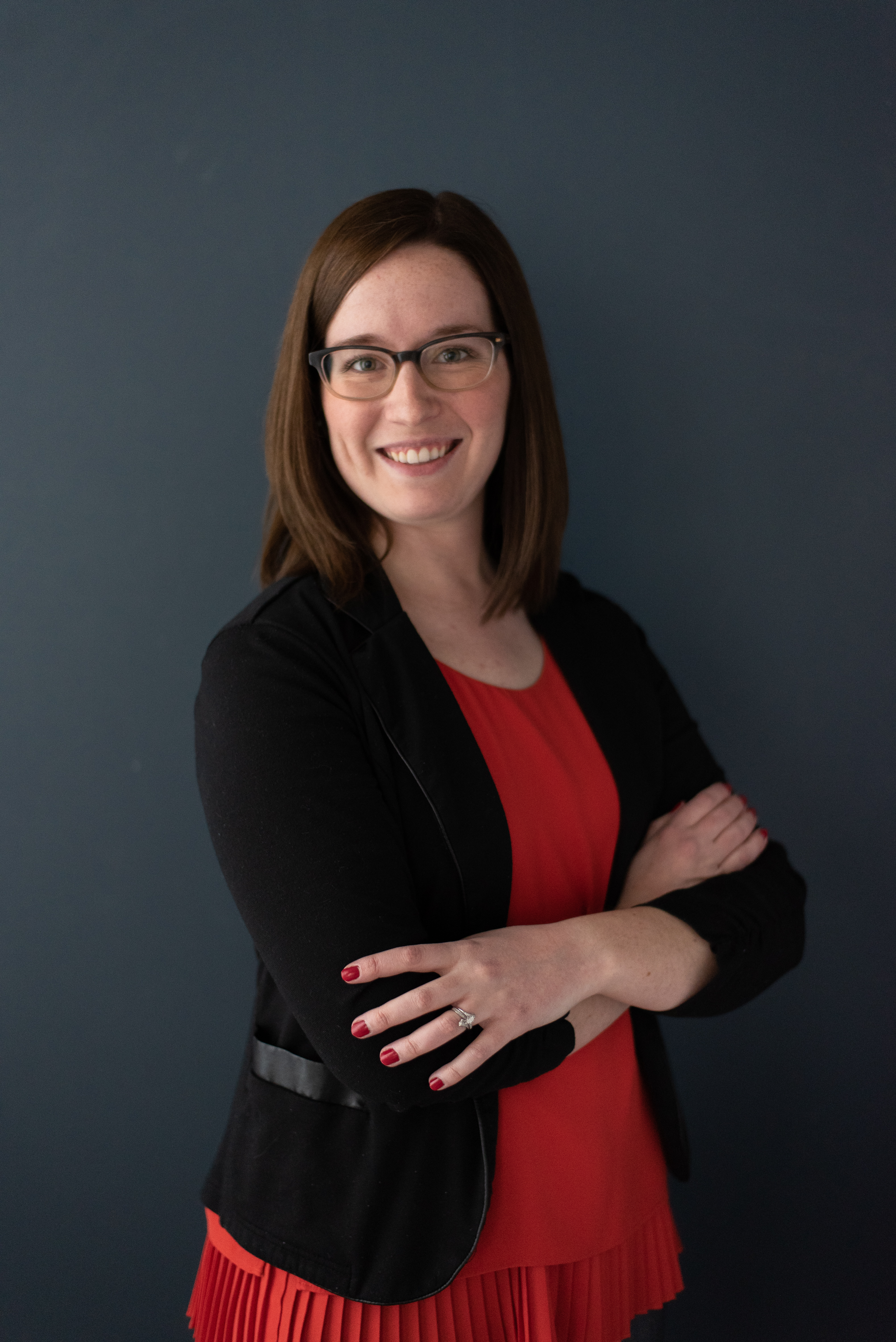 Portrait of Caroline in front of a dark background. In the photo, she has shoulder-length dark brown hair, brown-rimmed glasses, and red-painted nails. She's smiling and wearing a bright reddish-orange blouse with a black blazer.